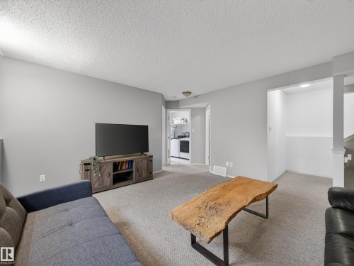 The property features a spacious room with light-colored walls and carpeting, a television console, and a clear view into a functional kitchen area - 1665 Tompkins Place, Edmonton, AB - Indoor Photo Showing Living Room