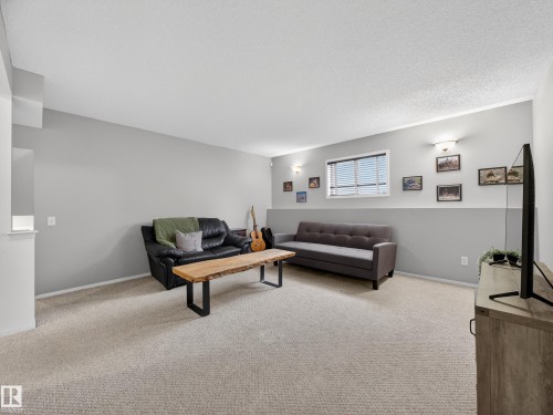 The property features a bright living area with light-colored carpeting, a window with blinds, and wall-mounted light fixtures - 1665 Tompkins Place, Edmonton, AB - Indoor
