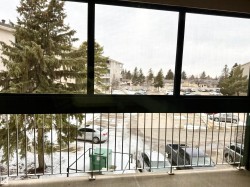 The property features a balcony with a metal railing, overlooking a snow-covered parking area and mature evergreen trees - 