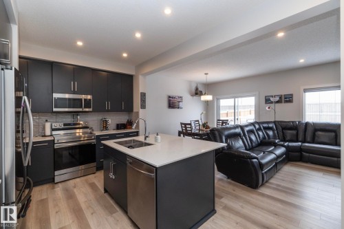 The open concept living area features light-toned flooring, recessed lighting, and a kitchen with dark cabinetry, stainless steel appliances, and a central island with a sink - 159 Larch Crescent, Leduc, AB - Indoor Photo Showing Kitchen With Double Sink