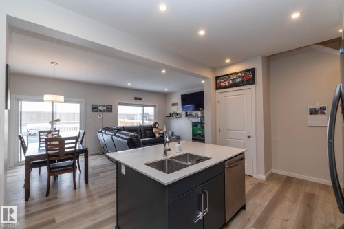 The kitchen features a central island with a sink and a stainless steel dishwasher, complemented by light-colored flooring throughout the open concept space - 159 Larch Crescent, Leduc, AB - Indoor Photo Showing Kitchen With Double Sink
