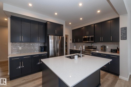 Modern kitchen featuring dark cabinetry, stainless steel appliances, a white quartz countertop island with an integrated sink, and light wood flooring - 159 Larch Crescent, Leduc, AB - Indoor Photo Showing Kitchen With Double Sink With Upgraded Kitchen