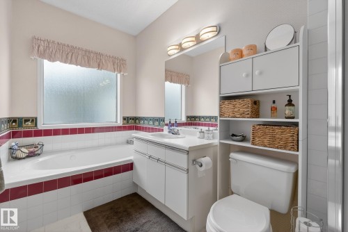 The bathroom features a built-in bathtub with white and red accent tiling, a vanity with a white countertop and double-door cabinets, and a toilet with an open shelving unit - 17204 113 Street, Edmonton, AB - Indoor Photo Showing Bathroom
