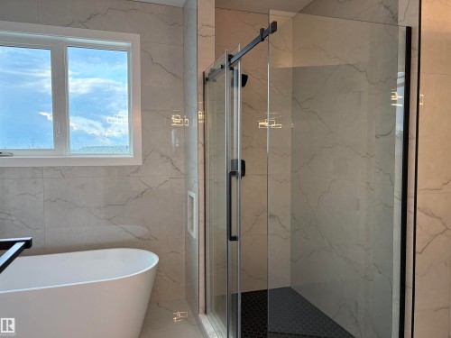 Freestanding soaking tub with a black caddy, glass-enclosed shower with black hexagonal tile flooring, and white tiled walls - 1039 151 Ave, Edmonton, AB 