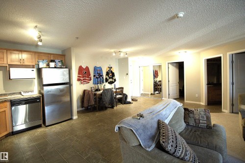 Open concept living area featuring a kitchen with stainless steel appliances, wood cabinetry, and a stone-look countertop, alongside a carpeted lounge area - 1204 330 Clareview Station Drive, Edmonton, AB - Indoor Photo Showing Kitchen
