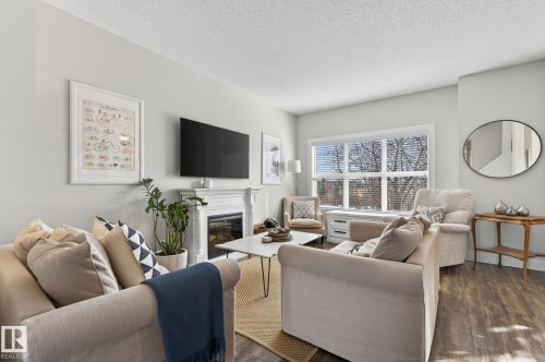 Living room featuring a fireplace with a white mantel, a large window with blinds, and wood-style flooring - 9084 Rosenthal Link, Edmonton, AB - Indoor Photo Showing Living Room With Fireplace
