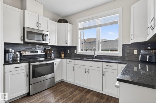 The kitchen features white cabinetry, dark countertops, a dark tile backsplash, stainless steel appliances, and a window above the sink - 9084 Rosenthal Link, Edmonton, AB - Indoor Photo Showing Kitchen