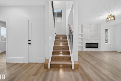 The property features light-toned flooring throughout, a staircase with individual step lighting, and a living area with a floor-to-ceiling tiled fireplace and built-in shelving - 6117 Crawford Drive, Edmonton, AB 