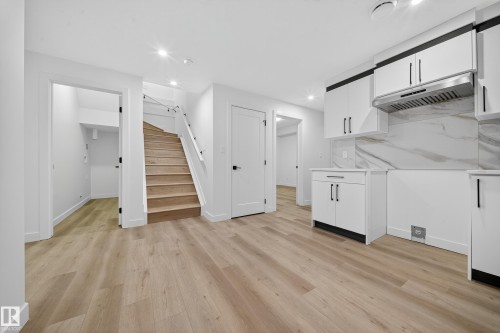 Open concept space featuring light wood flooring, a modern kitchen area with white cabinetry, dark contrasting handles, and a marble-style backsplash - 6117 Crawford Drive, Edmonton, AB 