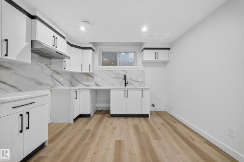 The kitchen features white cabinetry with black hardware, a contrasting black accent trim, white countertops, and a white backsplash with grey and gold veining - 6117 Crawford Drive, Edmonton, AB 