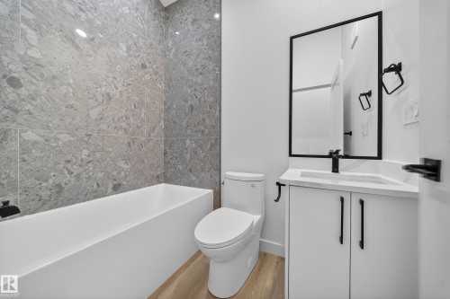 The bathroom features a modern vanity with a rectangular sink, a black-framed mirror, a bathtub with grey tile surround, and light wood-look flooring - 6117 Crawford Drive, Edmonton, AB 
