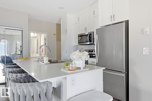 The kitchen features a large island with an integrated sink and an extendable faucet, white cabinetry, a stainless steel microwave, and a stainless steel refrigerator - 155 1818 Rutherford Road, Edmonton, AB - Indoor Photo Showing Kitchen With Stainless Steel Kitchen With Upgraded Kitchen