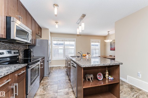 The kitchen features stainless steel appliances, granite countertops, and rich wood cabinetry, complemented by a mosaic tile backsplash - 31 4029 Orchards Drive, Edmonton, autreother - Indoor Photo Showing Kitchen With Upgraded Kitchen