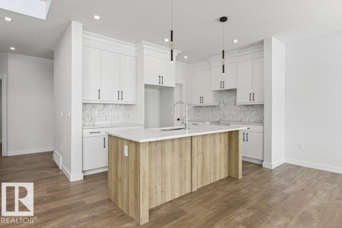 The kitchen features white cabinetry, a light-colored kitchen island with a sink, pendant lighting, and a white backsplash with a patterned design - 5018 53 Avenue, Calmar, AB - Indoor Photo Showing Kitchen With Upgraded Kitchen