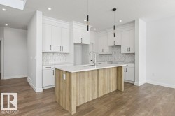 The kitchen features white cabinetry, a light-colored kitchen island with a sink, pendant lighting, and a white backsplash with a patterned design - 