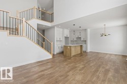 Open-concept living space featuring light-toned flooring, a two-story staircase with light wood and black metal railings, and a kitchen with white cabinetry and an island - 