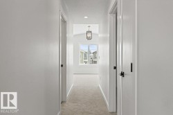 Hallway with light-colored walls and carpeting, featuring several doors with dark hardware, and a window providing natural light at the far end - 