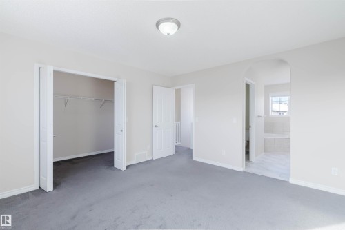 This room features soft gray carpeting, a closet with bi-fold doors and wire shelving, and an arched entryway leading to a bathroom with tiled flooring and a window - 1135 112 Street, Edmonton, AB - Indoor Photo Showing Other Room