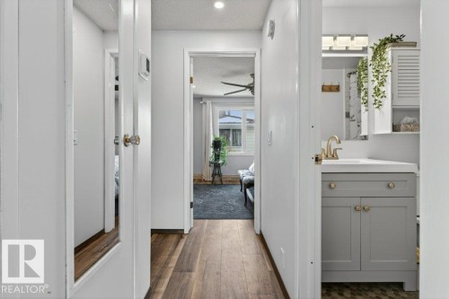 This interior hallway features wood-style flooring and provides access to a room with a ceiling fan and a bathroom with a vanity, sink, and mirror - 2655 89 Street, Edmonton, AB - Indoor