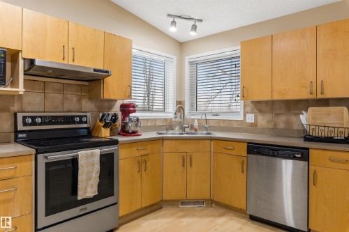 Kitchen featuring light wood cabinetry, stainless steel appliances, a double window with horizontal blinds, and a tiled backsplash - 1233 Ormsby Lane, Edmonton, AB - Indoor Photo Showing Kitchen With Double Sink
