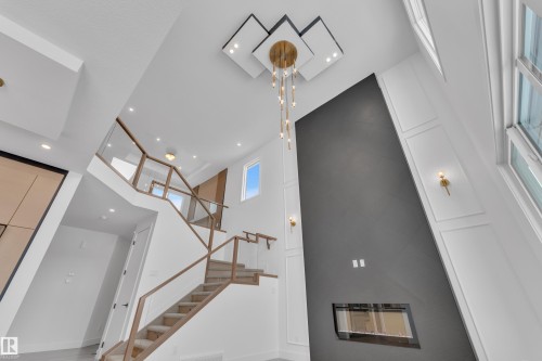 The property features a grand staircase with glass railings and wood handrails, an accent wall with a modern fireplace, and a striking chandelier - 503 Creekside Bay, Leduc, AB - Indoor Photo Showing Other Room