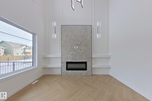 The property features a spacious room with light-colored flooring arranged in a herringbone pattern, a wide window, and a fireplace with a stone surround - 8425 Mayday Link Link, Edmonton, AB - Indoor With Fireplace