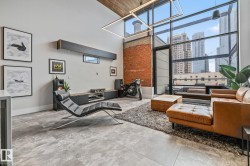 The living space features high ceilings, exposed brick, and large windows that provide abundant natural light and views of city buildings - 