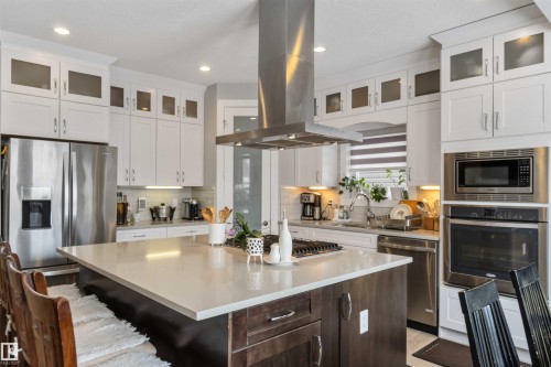 The kitchen features a spacious island with a light-colored countertop, white upper cabinetry, and stainless steel appliances including a refrigerator, oven, microwave, and dishwasher - 4 Lakeshore Cove, Beaumont, AB 
