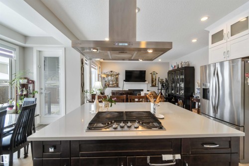 The kitchen features a large island with a built-in cooktop and a range hood overhead, white cabinetry, and stainless steel appliances - 4 Lakeshore Cove, Beaumont, AB 