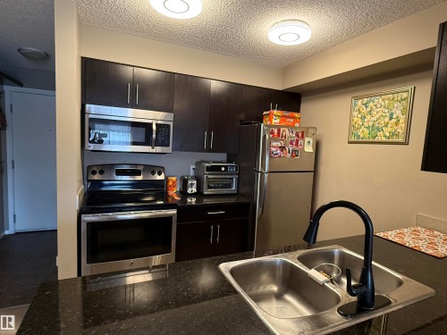 The kitchen features dark cabinetry, stainless steel appliances including a microwave, oven, and refrigerator, and a dark stone countertop with a double basin sink and a black faucet - 308 25 Element Dr Drive N, St. Albert, AB - Indoor Photo Showing Kitchen With Stainless Steel Kitchen With Double Sink