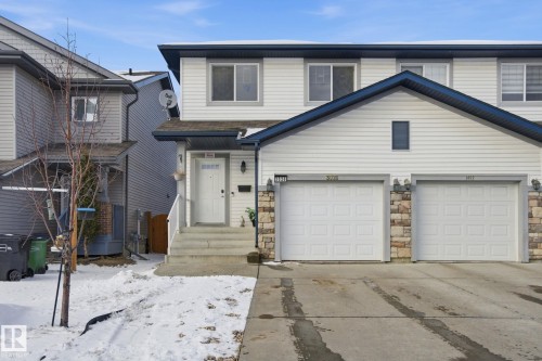The property features a white exterior with dark blue trim, a paved driveway, and two white garage doors with stone accents - 3026 16A Avenue, Edmonton, AB - Outdoor With Facade