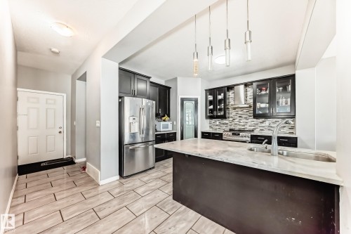 The kitchen features dark cabinetry, stainless steel appliances, a tile backsplash, and an island with a sink and countertop space - 16720 61 Street, Edmonton, AB - Indoor Photo Showing Kitchen With Upgraded Kitchen