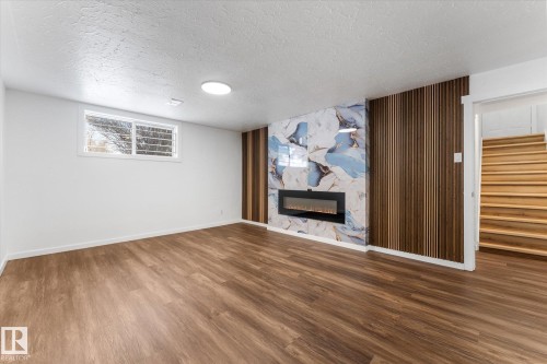 The property features a spacious room with wood-look flooring, a modern electric fireplace set into a decorative marble-patterned wall, and an accent wall with vertical wood paneling - 13116 128A Avenue, Edmonton, AB - Indoor With Fireplace