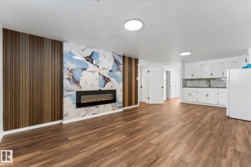 The property features a spacious room with wood-look flooring, an electric fireplace with a decorative stone wall surround, and a kitchen area with white cabinetry and a sink - 13116 128A Avenue, Edmonton, AB - Indoor Photo Showing Living Room With Fireplace