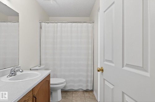 The bathroom features a vanity with a white countertop and an integrated sink, a toilet, and a shower area with a white shower curtain - 22 Riverpointe, Fort Saskatchewan, AB - Indoor Photo Showing Bathroom