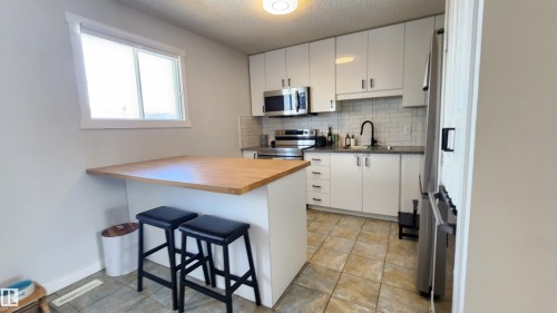 The kitchen features a dining island with a wooden countertop, white cabinetry, stainless steel appliances, and tiled flooring - 8326 157 Avenue, Edmonton, AB - Indoor Photo Showing Kitchen
