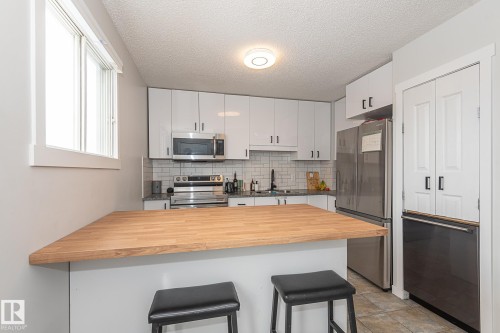 The kitchen features white cabinetry, a light wood-toned breakfast bar, and stainless steel appliances - 8326 157 Avenue, Edmonton, AB - Indoor Photo Showing Kitchen