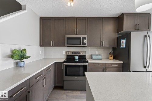 The kitchen features dark wood cabinetry, light countertops, stainless steel appliances including a refrigerator, microwave, and oven with an electric stovetop, and a tiled backsplash - 8225 24 Avenue, Edmonton, AB - Indoor Photo Showing Kitchen