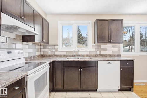 The kitchen features dark wood cabinetry, a subway tile backsplash, a double basin sink, and white appliances - 6 Garden Crescent, St. Albert, AB - Indoor Photo Showing Kitchen With Double Sink