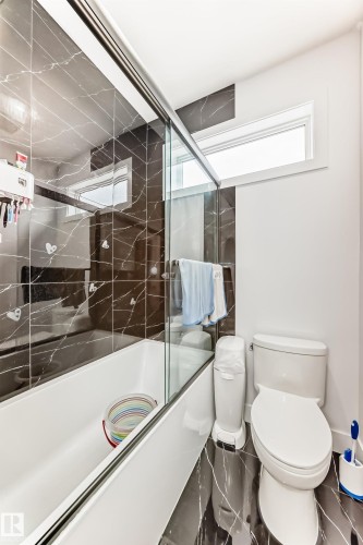 The bathroom features a bathtub and shower combination with a glass enclosure, dark marble-effect wall and floor tiles, and a window providing natural light - 15108 11 Street, Edmonton, AB - Indoor Photo Showing Bathroom