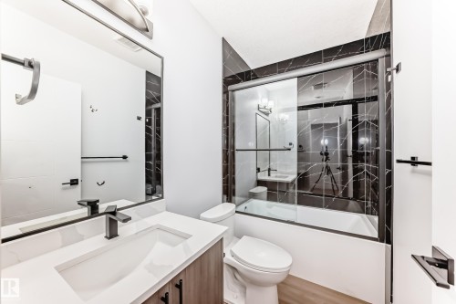 Bathroom featuring a vanity with a white countertop and dark faucet, a large mirror, and a shower-tub combination with dark tiled walls and a sliding glass door - 15108 11 Street, Edmonton, AB - Indoor Photo Showing Bathroom