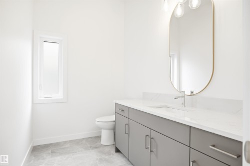 Well-appointed bathroom featuring a vanity with a white countertop and gray cabinetry, a gold-framed oval mirror, and a window providing natural light - 9807 90 Avenue, Edmonton, AB - Indoor Photo Showing Bathroom