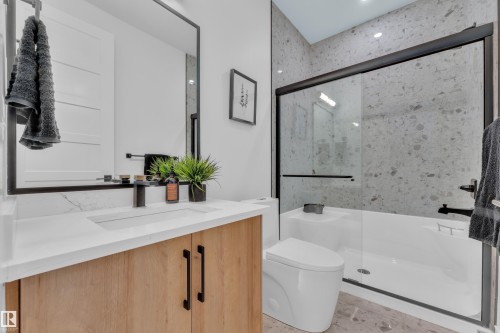 Bathroom featuring a light wood vanity with a white countertop and rectangular sink, a black framed mirror, and a shower-tub combination with a glass enclosure and gray tiled walls - 110 Eldridge Point(E), St. Albert, AB - Indoor Photo Showing Bathroom