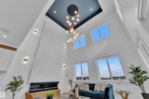 The living area features a high ceiling with a contemporary chandelier, a white textured accent wall, and multiple windows providing natural light - 110 Eldridge Point(E), St. Albert, AB - Indoor With Fireplace