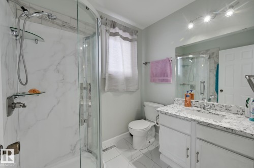 The bathroom features a curved glass shower enclosure with a marble-patterned surround, a white vanity with a patterned countertop, and a mirror with track lighting above - 8507 152B Avenue, Edmonton, AB - Indoor Photo Showing Bathroom