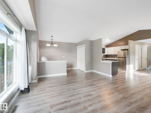 This open concept living space features light wood-style flooring, a large window with white curtains, and a kitchen area with stainless steel appliances and light cabinetry - 5035 51 Avenue, Lamont, AB - Indoor