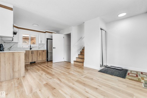The property features an open-concept living space with light wood flooring, a modern kitchen with white and wood cabinetry, and a stainless steel refrigerator - 10865 21 Ave, Edmonton, AB - Indoor Photo Showing Kitchen