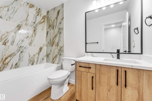 The bathroom features a modern vanity with a rectangular sink, a large mirror with integrated lighting, and a white bathtub with marble-look tiled surround - 10865 21 Ave, Edmonton, AB - Indoor Photo Showing Bathroom