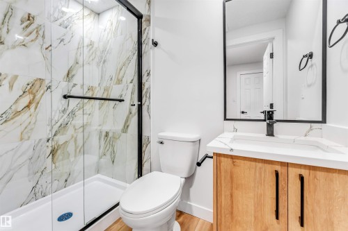 Bathroom featuring a glass-enclosed shower with marbled tile, a vanity with a light wood finish and a white countertop, and a mirror with a black frame - 10865 21 Ave, Edmonton, AB - Indoor Photo Showing Bathroom