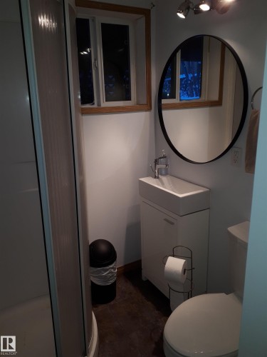 Compact bathroom featuring a shower stall with a clear door, a white vanity with an integrated sink, a round mirror, and a window with wood trim - 5512 50Th Ave, Wetaskiwin, AB - Indoor Photo Showing Bathroom